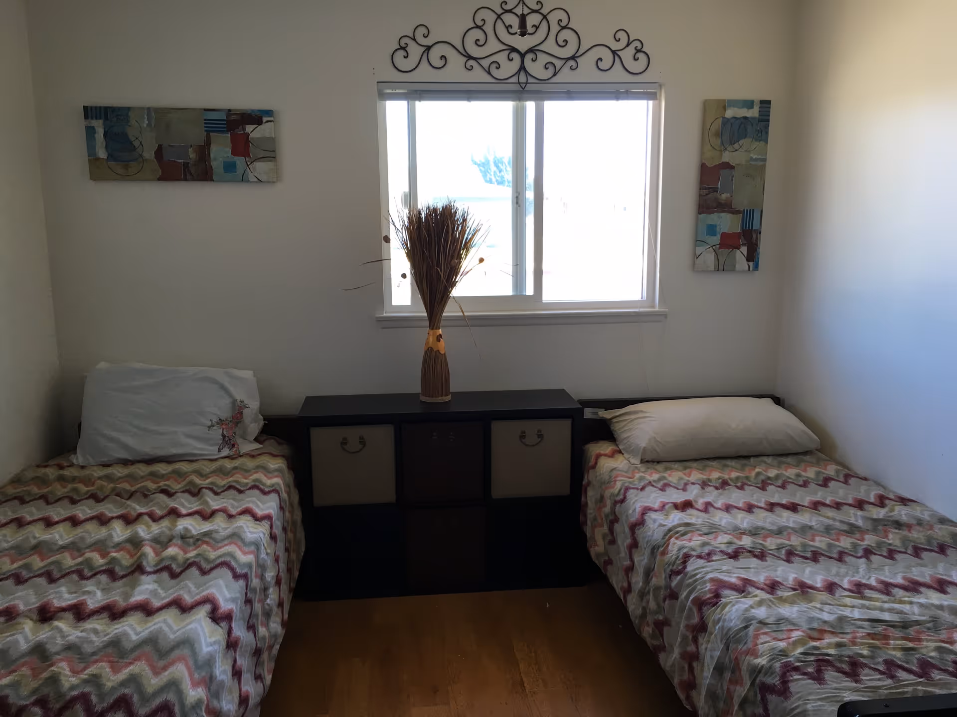 A small bedroom with two single beds placed on opposite sides of the room. Each bed has a pillow and a zigzag patterned bedspread in shades of red, green, and beige. Between the beds is a black storage unit with six cubbies, some containing fabric drawers. On top of the storage unit is a decorative vase with dried plants. Above the window, there is a decorative black wrought iron wall piece. Two abstract paintings hang on the walls beside the window.