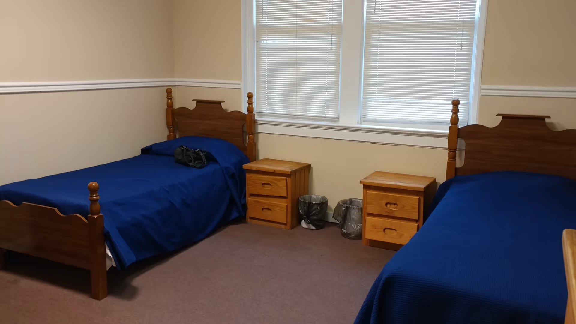 Two wooden twin beds with blue covers, nightstands, and windows in a beige bedroom.