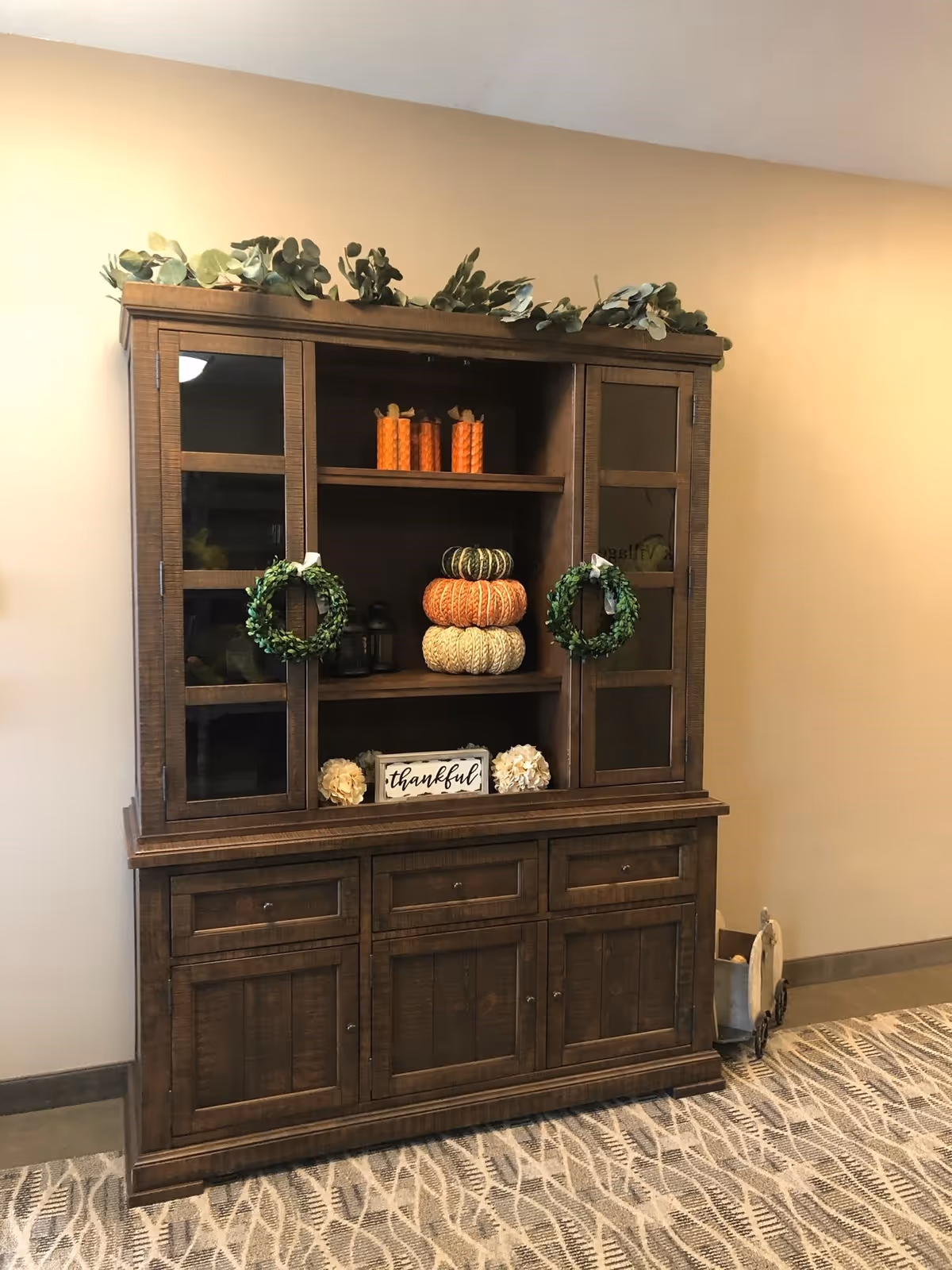 A wooden cabinet with glass doors on the upper section and closed drawers and cabinets on the lower section. The cabinet is decorated with two small green wreaths, three stacked decorative pumpkins in orange, green, and cream colors, two orange candles, two small lanterns, and a sign that reads 'thankful'. There is greenery on top of the cabinet and a patterned carpet on the floor.