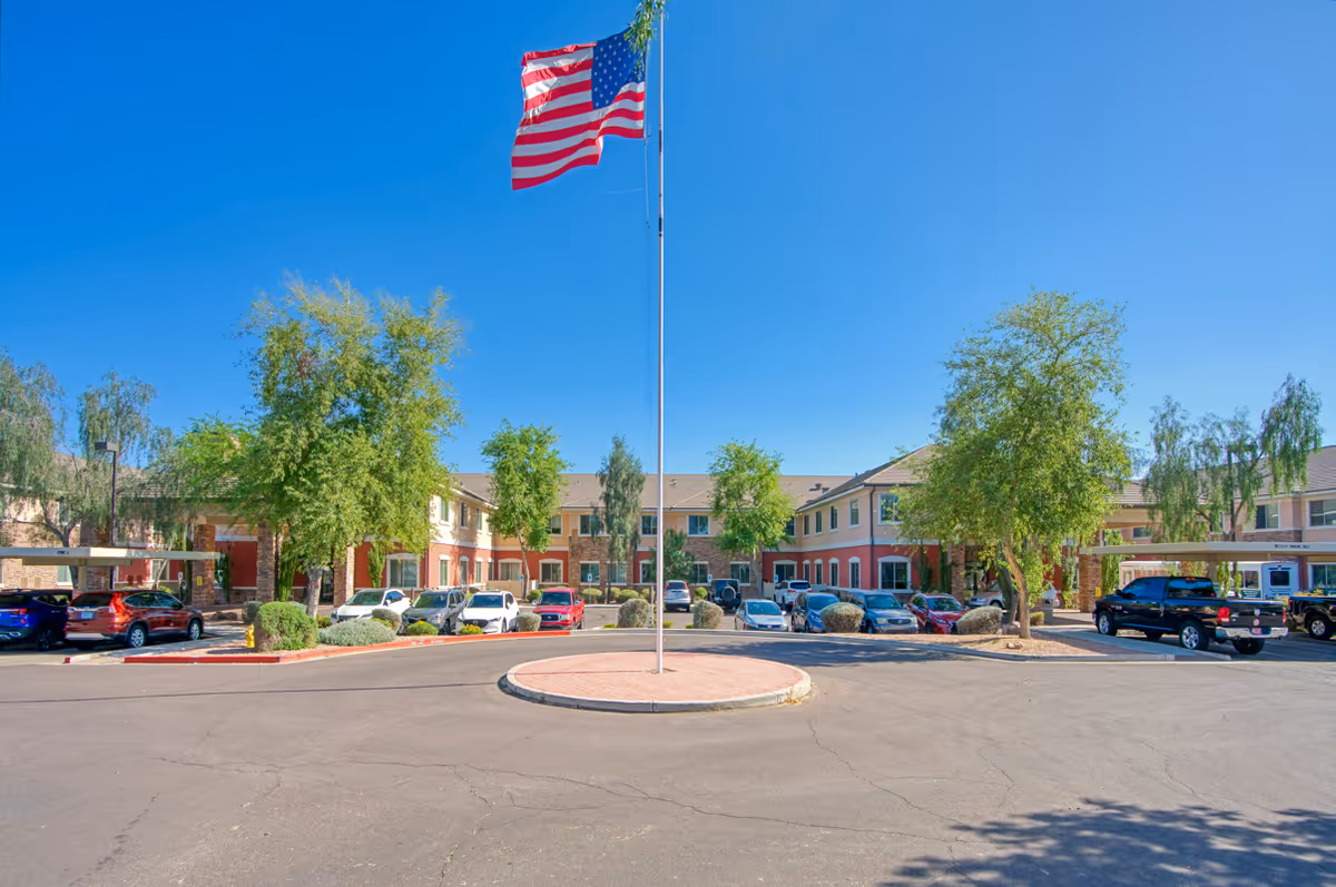 Front exterior view of Legacy House of Mesa, a two-story building with a parking lot in front. An American flag flies on a flagpole in the center of a circular driveway, surrounded by trees and parked cars under a clear blue sky.