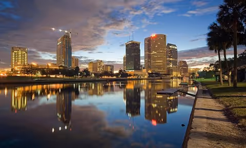 Sunset city skyline over calm water with tall buildings and a riverside walkway