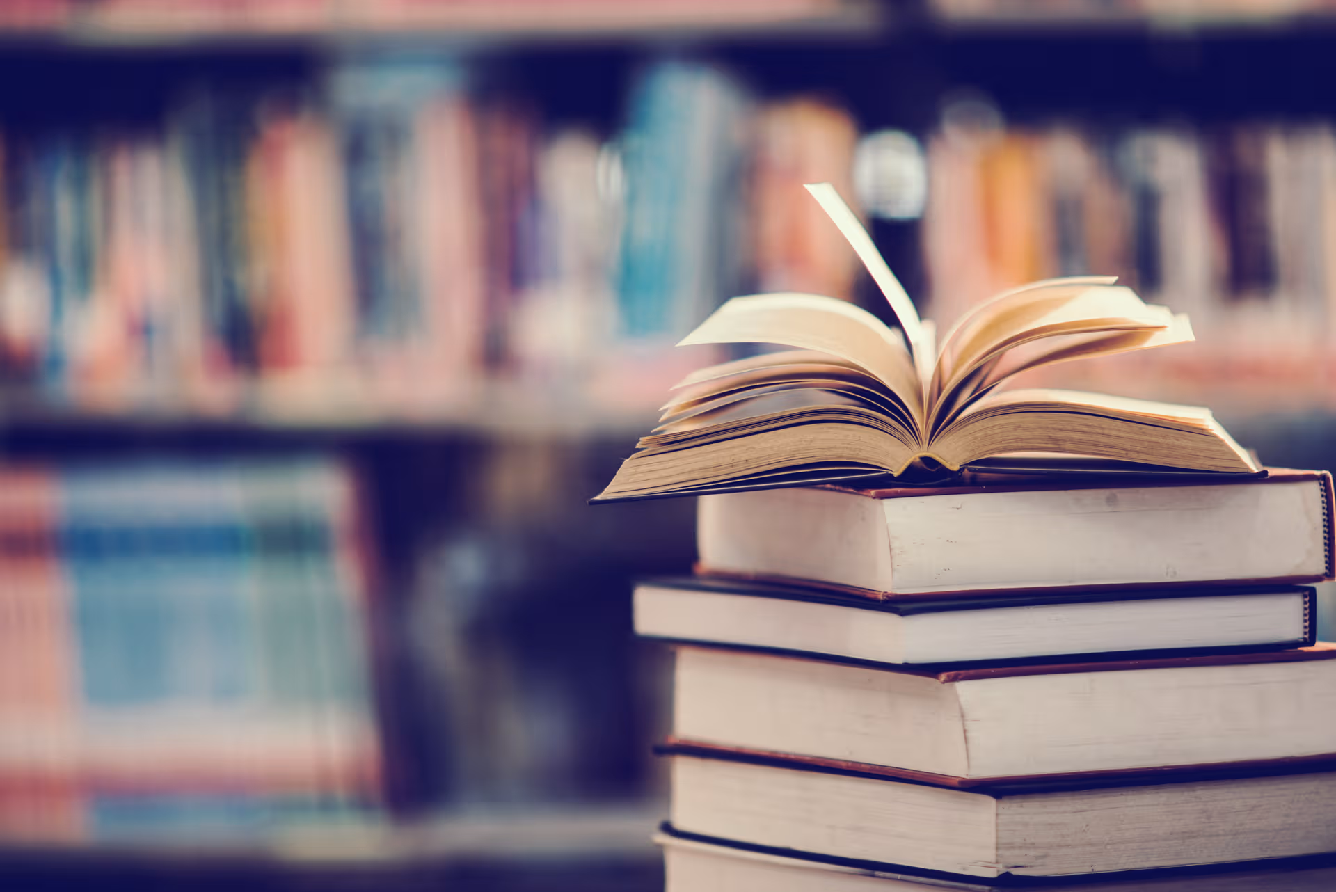 A stack of books with an open book on top against blurred library shelves.