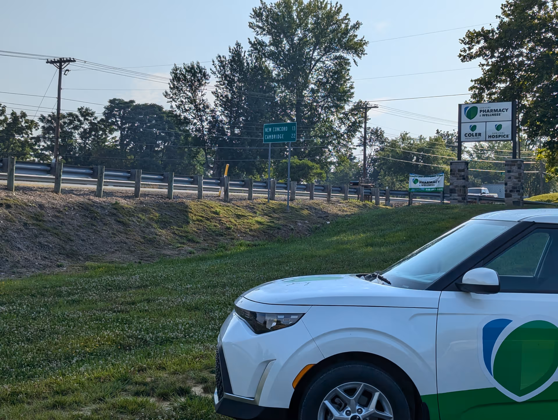 White vehicle with a green logo parked on a grassy roadside with signs in the background.