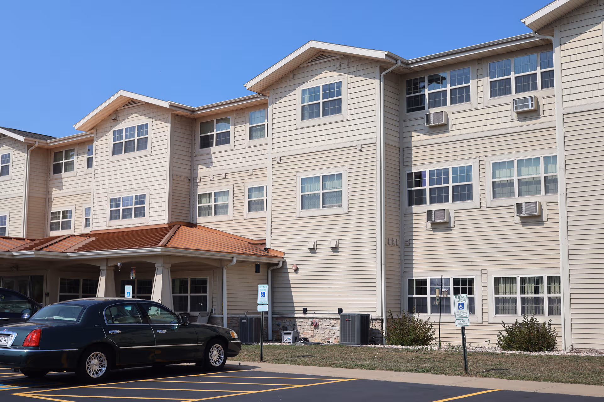 Three-story beige senior living building with windows, a covered entrance, and a parked sedan.