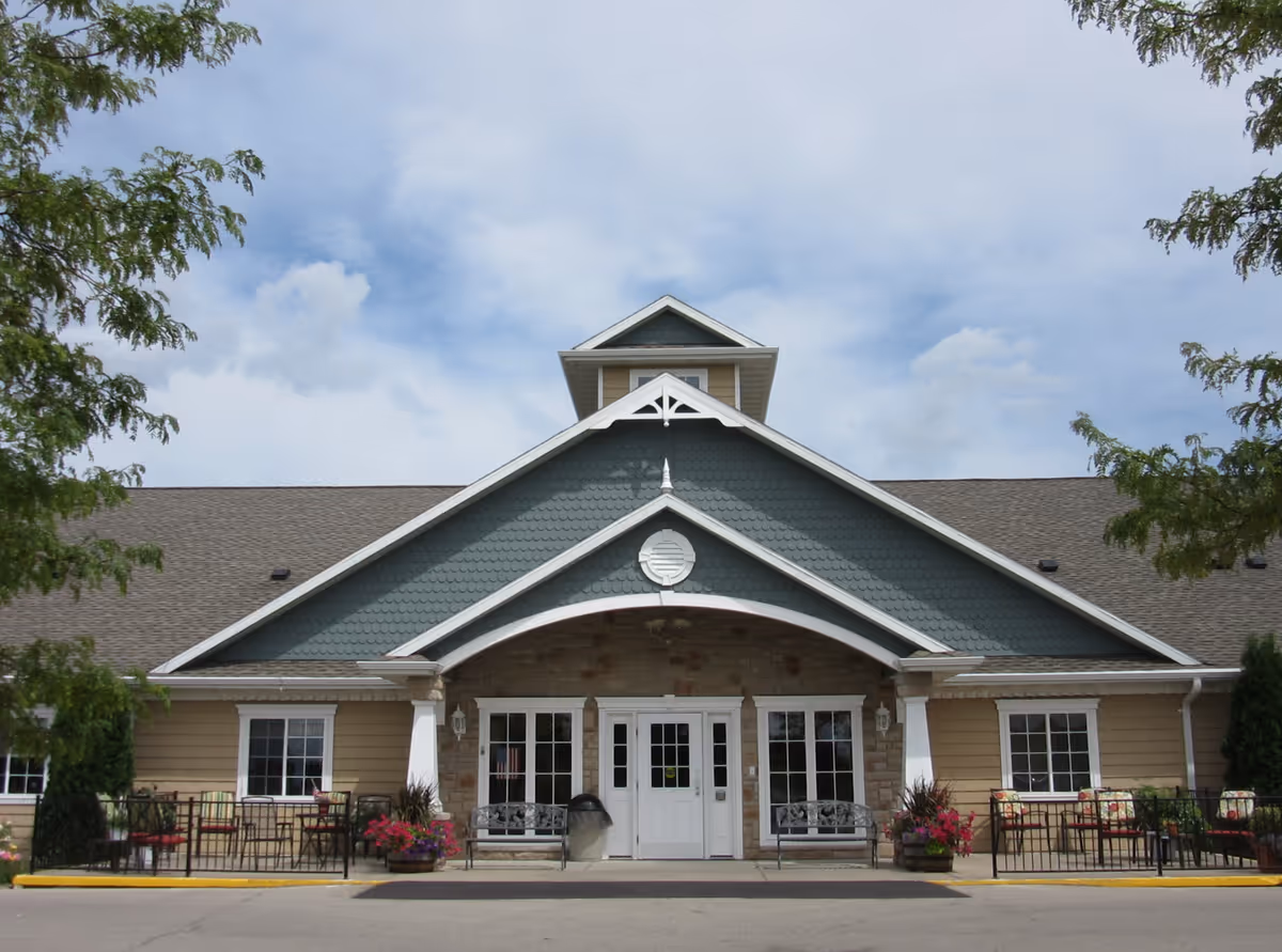 Front exterior view of a single-story building with a peaked roof and a small dormer window. The entrance has double white doors flanked by large windows and two benches on either side. There are flower pots with colorful flowers and outdoor seating areas with tables and chairs on both sides of the entrance. Trees frame the image on the left and right sides under a partly cloudy sky.