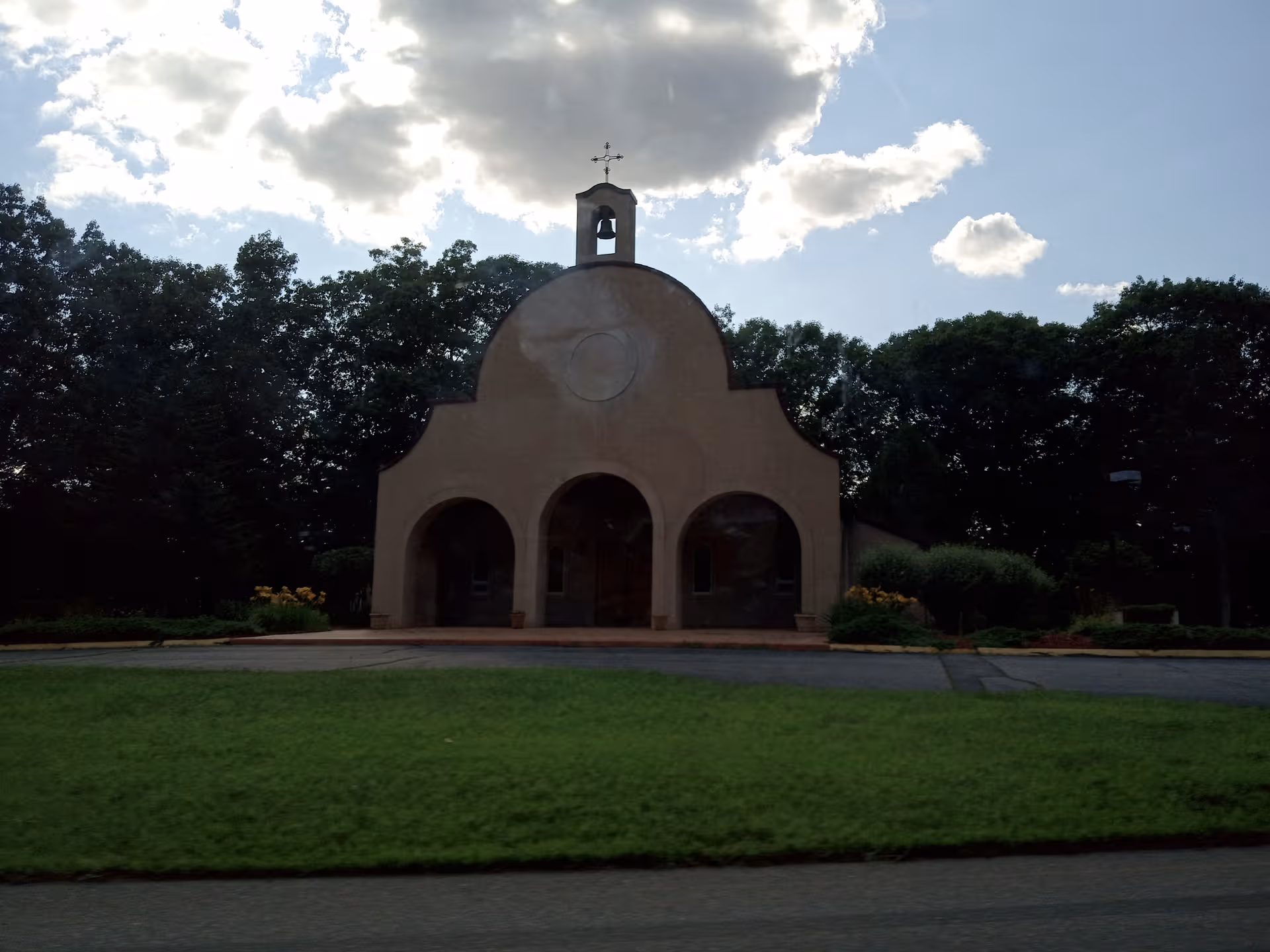 Front view of a small arched chapel with a cross, surrounded by trees and lawn.