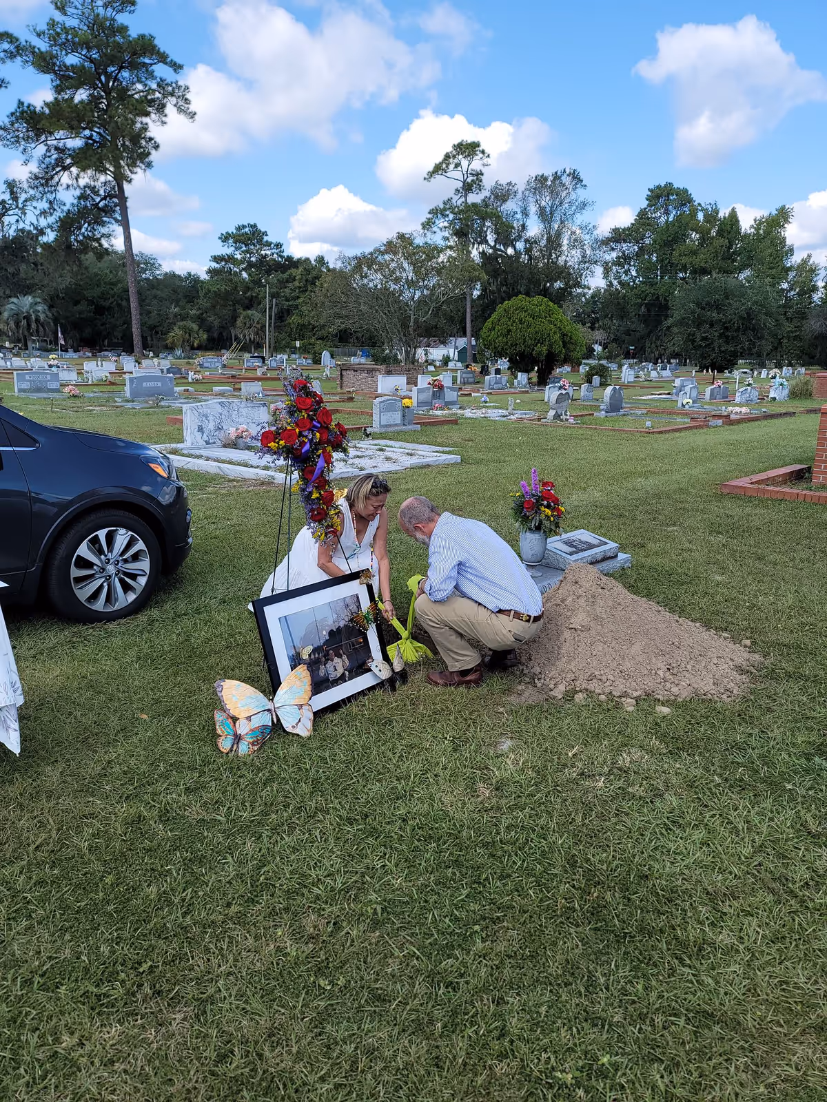 Two people kneel beside a gravestone in a cemetery with flowers and a framed photo.