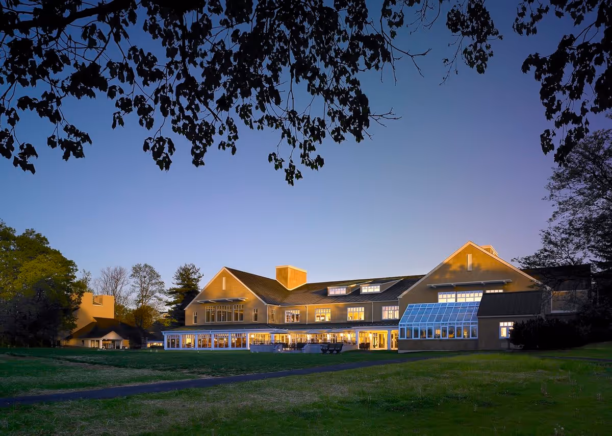 Exterior view of a large senior living facility building at dusk with lights glowing from the windows, surrounded by trees and a grassy area in the foreground.
