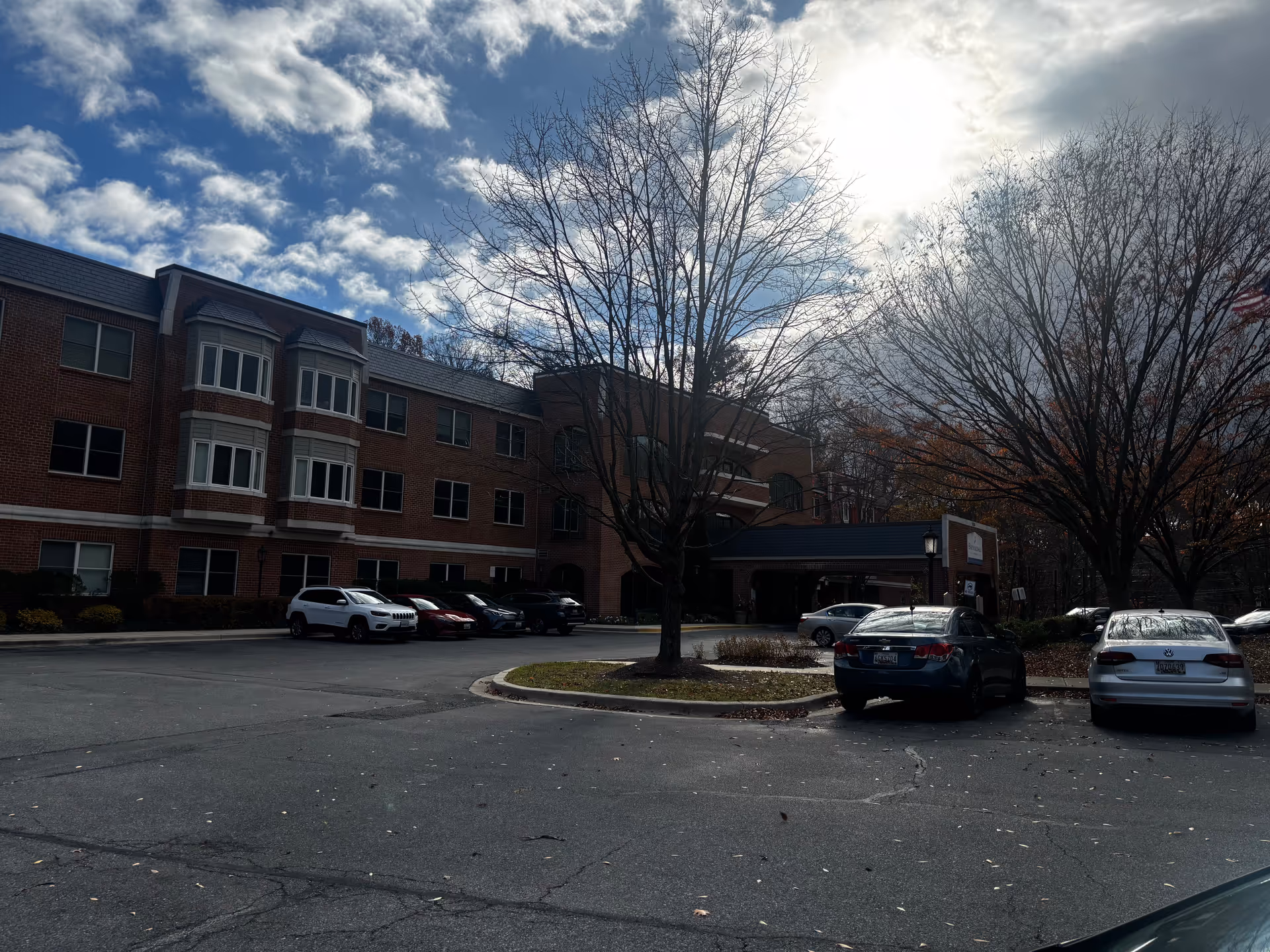 Front view of a three-story brick senior living building with a parking lot, several cars, and bare trees under a cloudy sky.