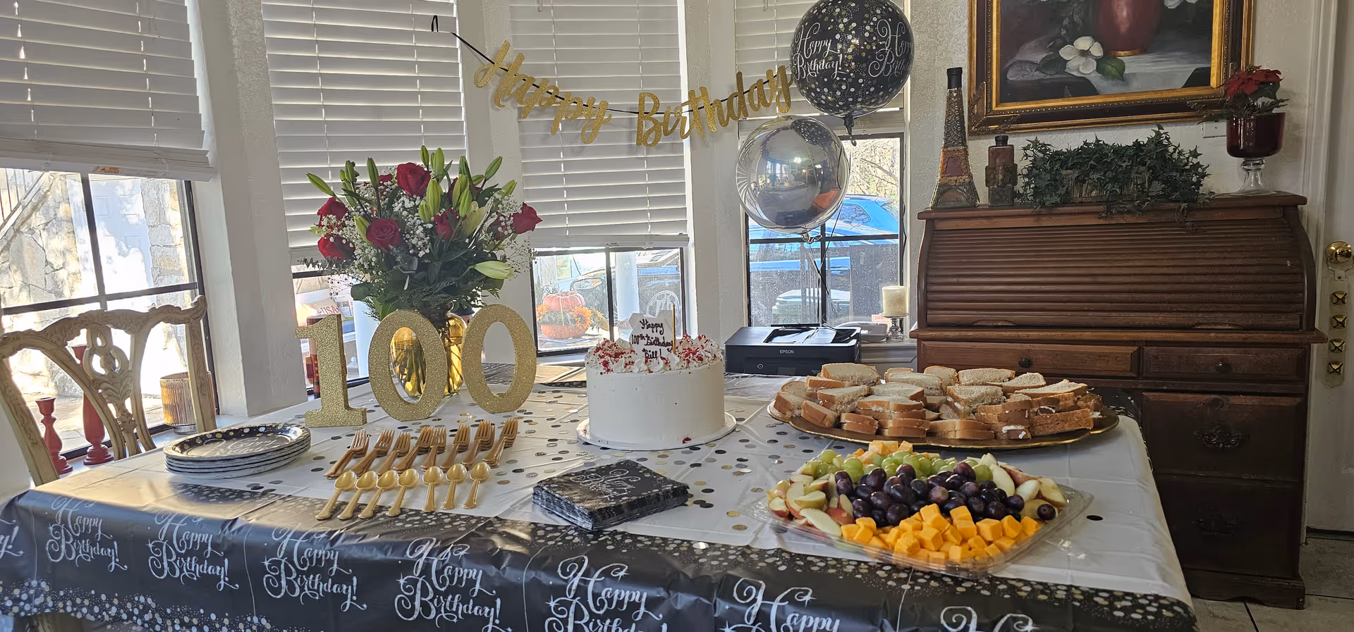 Indoor birthday setup with cake, fruit platter, balloons, flowers, and a Happy Birthday banner.