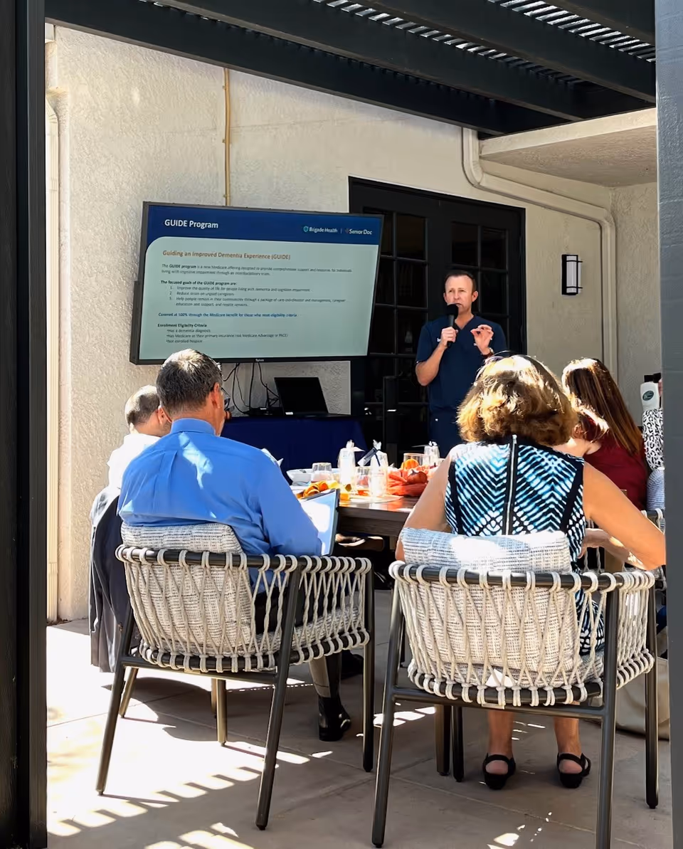 A presenter speaks into a microphone to a seated group at an outdoor patio.