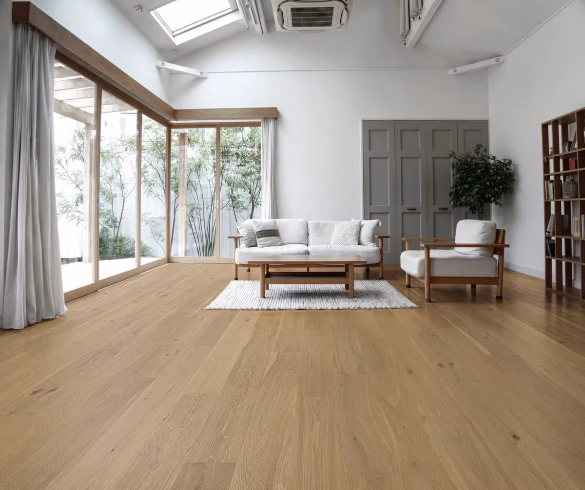 Bright, minimalist living room with cream furniture, wooden floors, large windows, and a bookshelf.