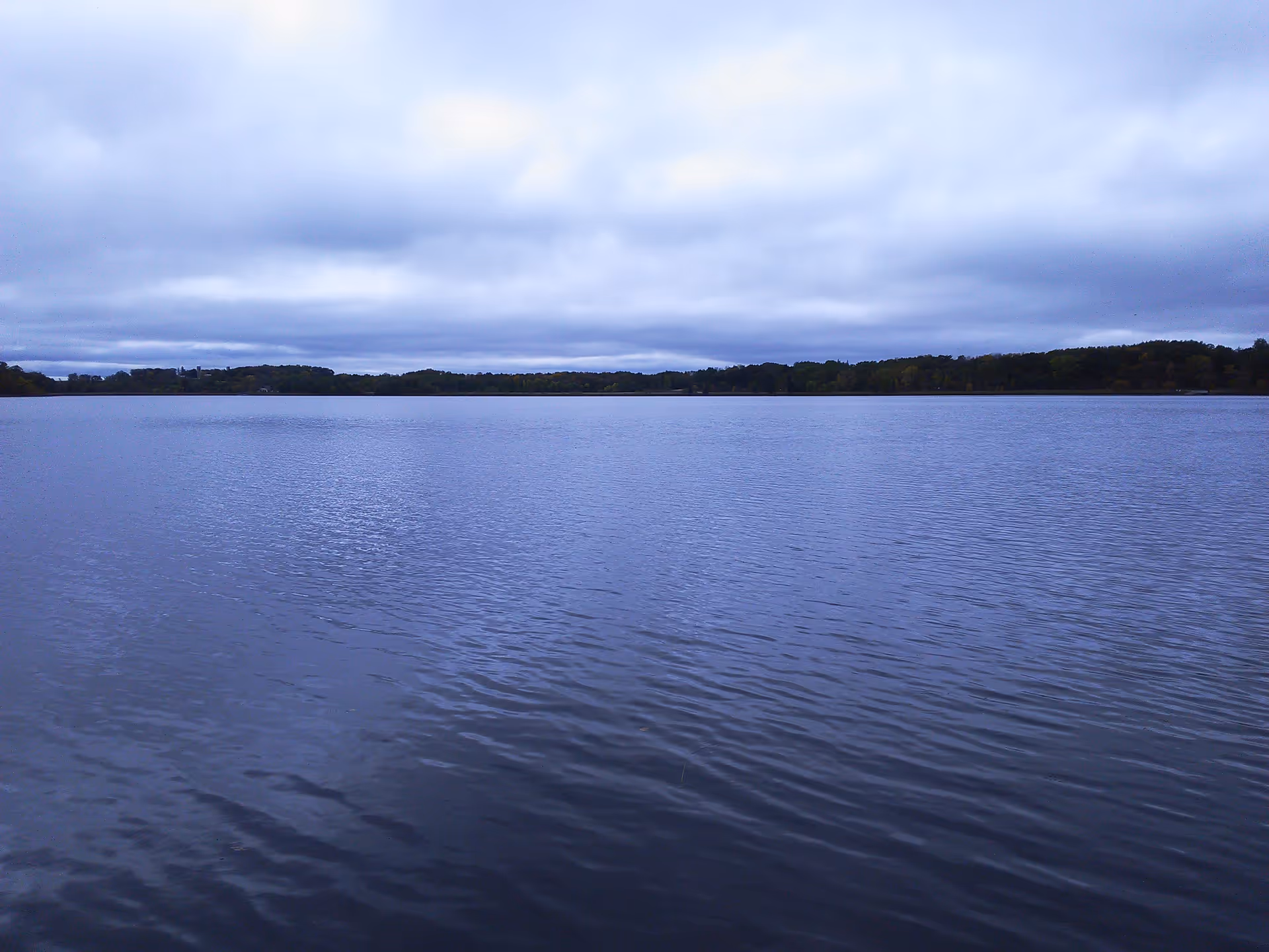 Calm lake stretches to tree-lined horizon beneath a cloudy, blue-tinged sky.