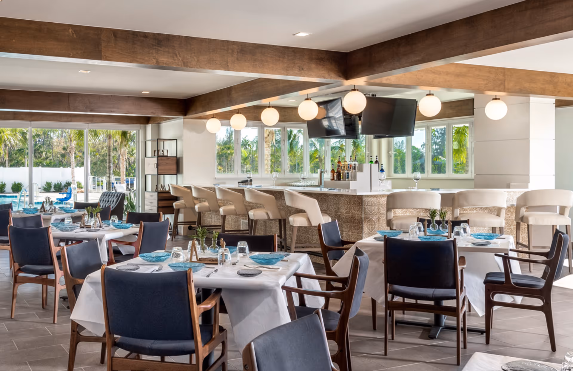 A bright and modern dining area with tables covered in white tablecloths, set with blue plates, glasses, and silverware. The room features dark wooden chairs with blue cushions, a bar with beige bar stools, hanging globe lights, and large windows showing greenery outside.