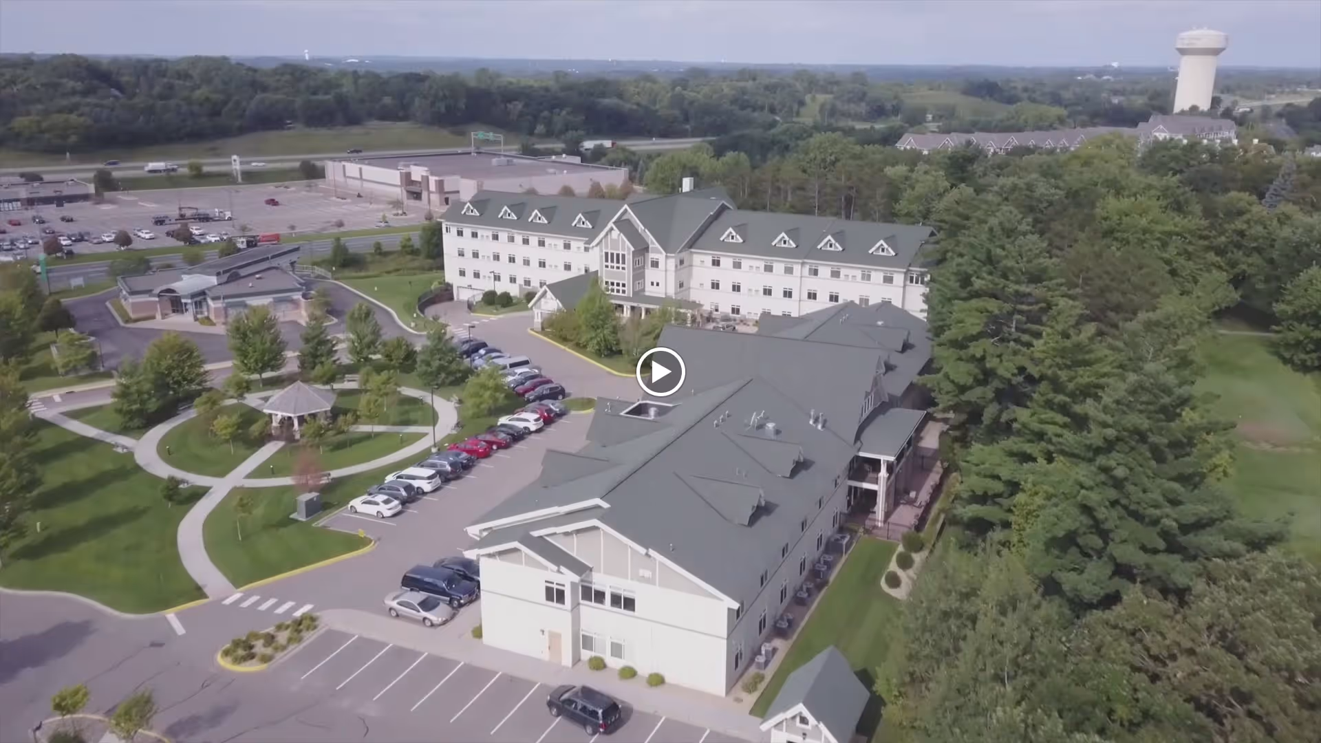 Aerial view of a senior living facility with multiple connected buildings surrounded by green lawns, trees, and walking paths. There is a gazebo in the garden area, several parked cars in the parking lot, and a water tower visible in the distance.