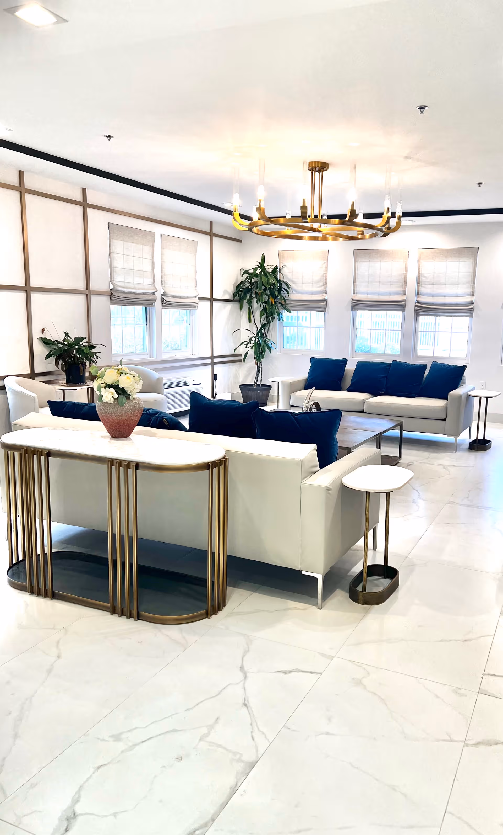 Bright modern lounge with white seating, blue cushions, greenery, and a gold-toned chandelier.