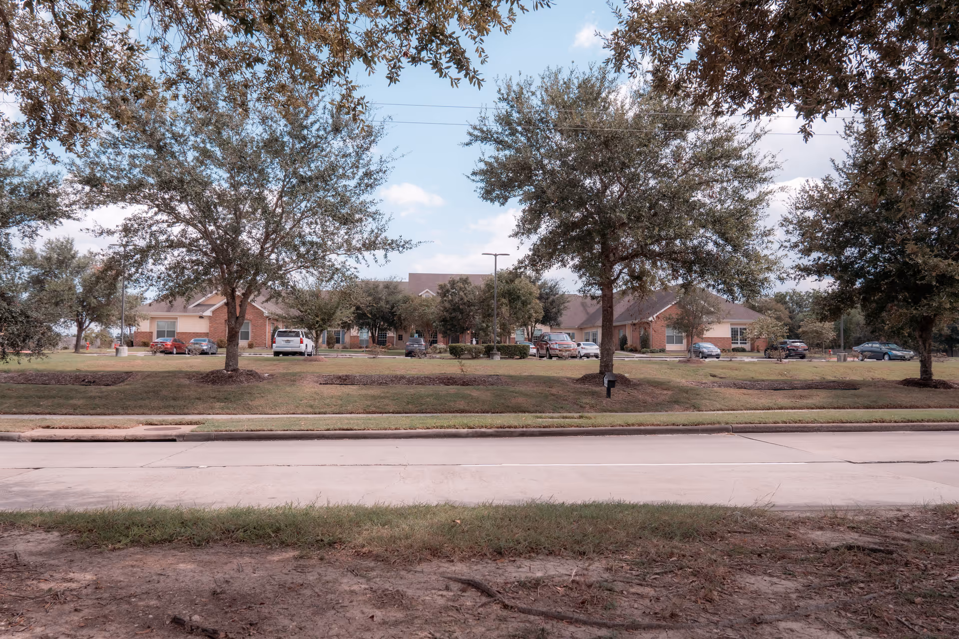 Front exterior view of a single-story brick building set behind a lawn, trees, and parked cars.