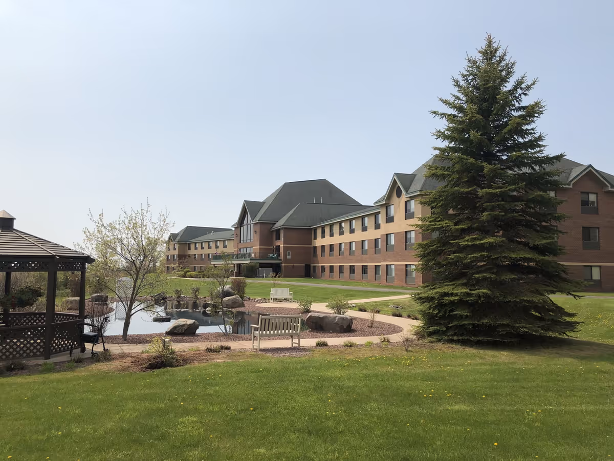 Exterior view of The Bluffs senior living building with landscaped grounds, a pond, gazebo, benches, and a large evergreen tree.