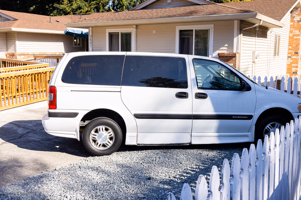 White van parked in driveway in front of a single-story home with a white picket fence.