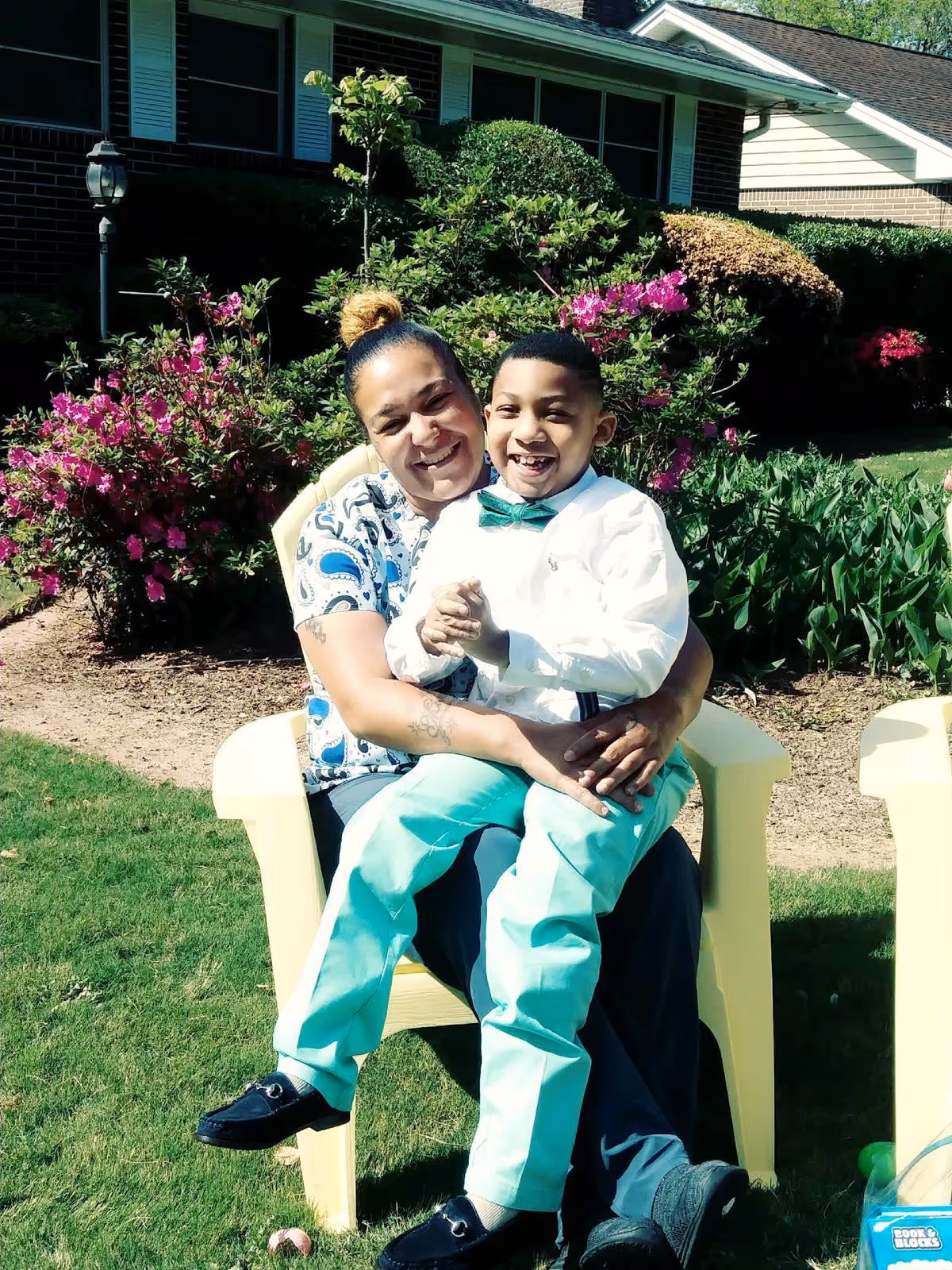 Woman and child smile while seated outdoors on yellow chairs beside a flowering garden.