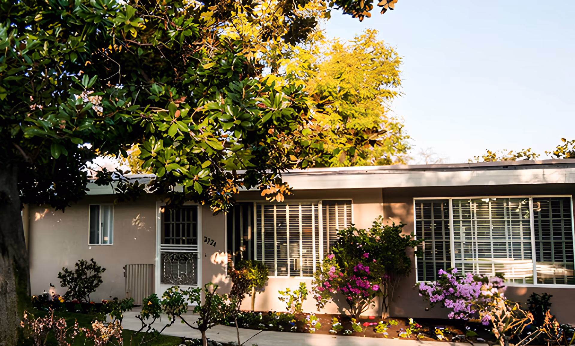 Exterior view of a single-story building with large windows and a white door, surrounded by green trees and colorful flowering plants in a garden.