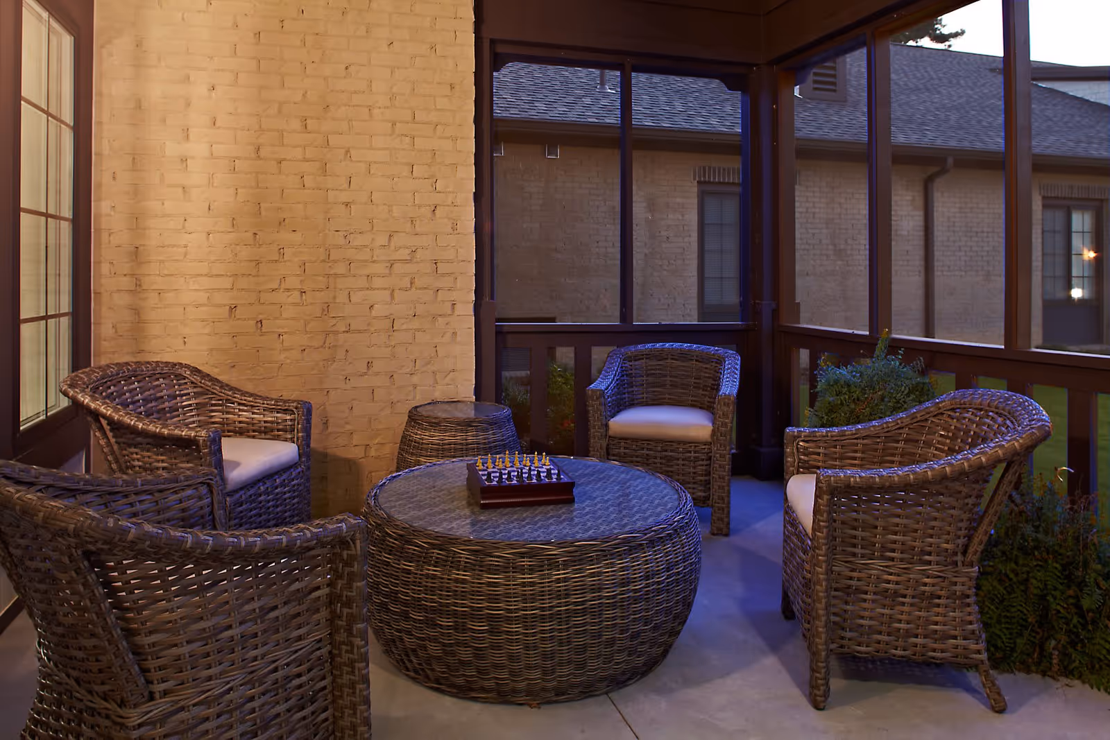 A cozy outdoor seating area with four wicker chairs with cushions arranged around a round wicker table with a glass top. A chessboard is placed on the table. The area is enclosed with screened windows and has a brick wall on one side.