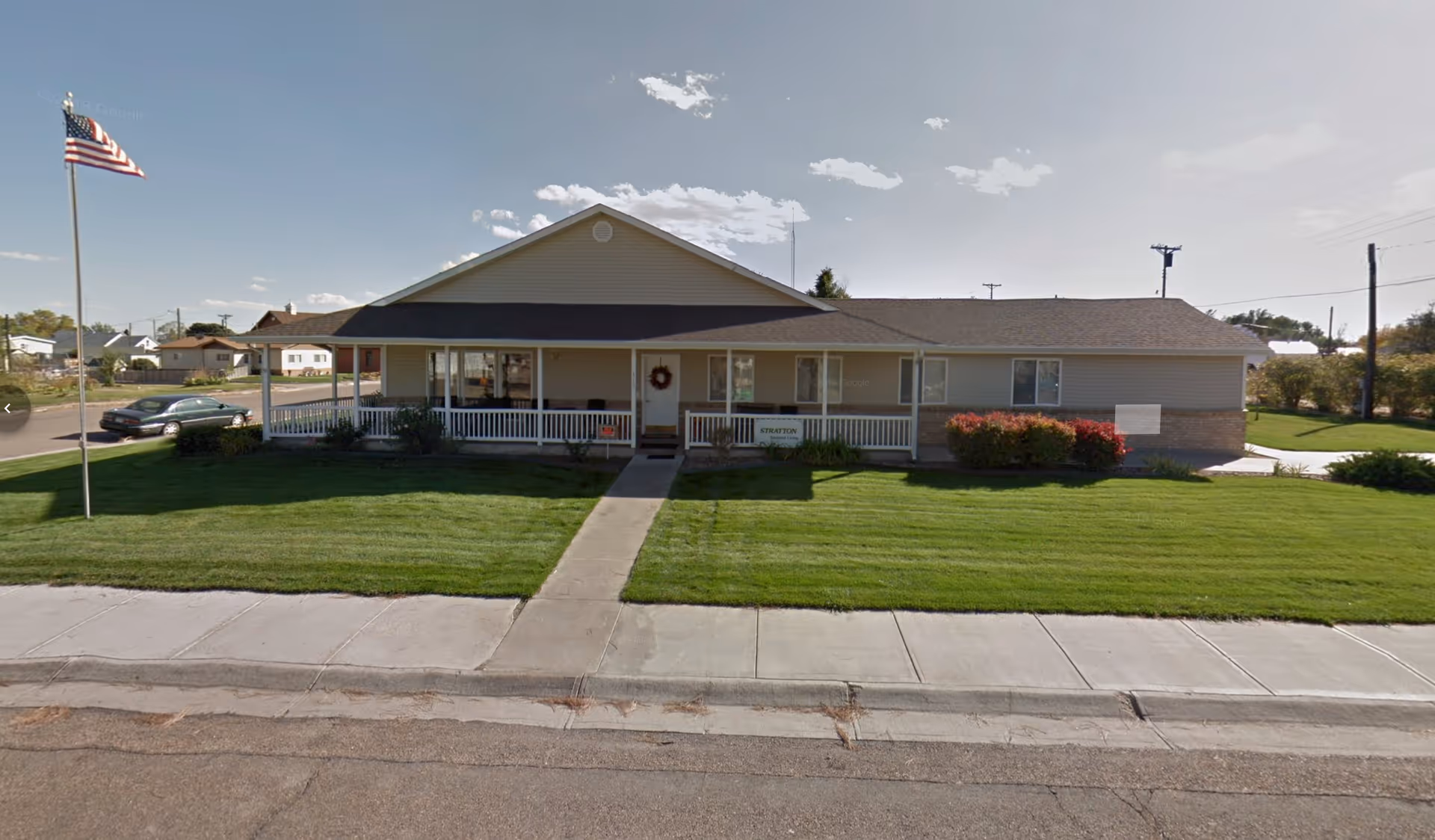 Single-story assisted living building with a covered front porch, walkway, manicured lawn and an American flag on a pole.