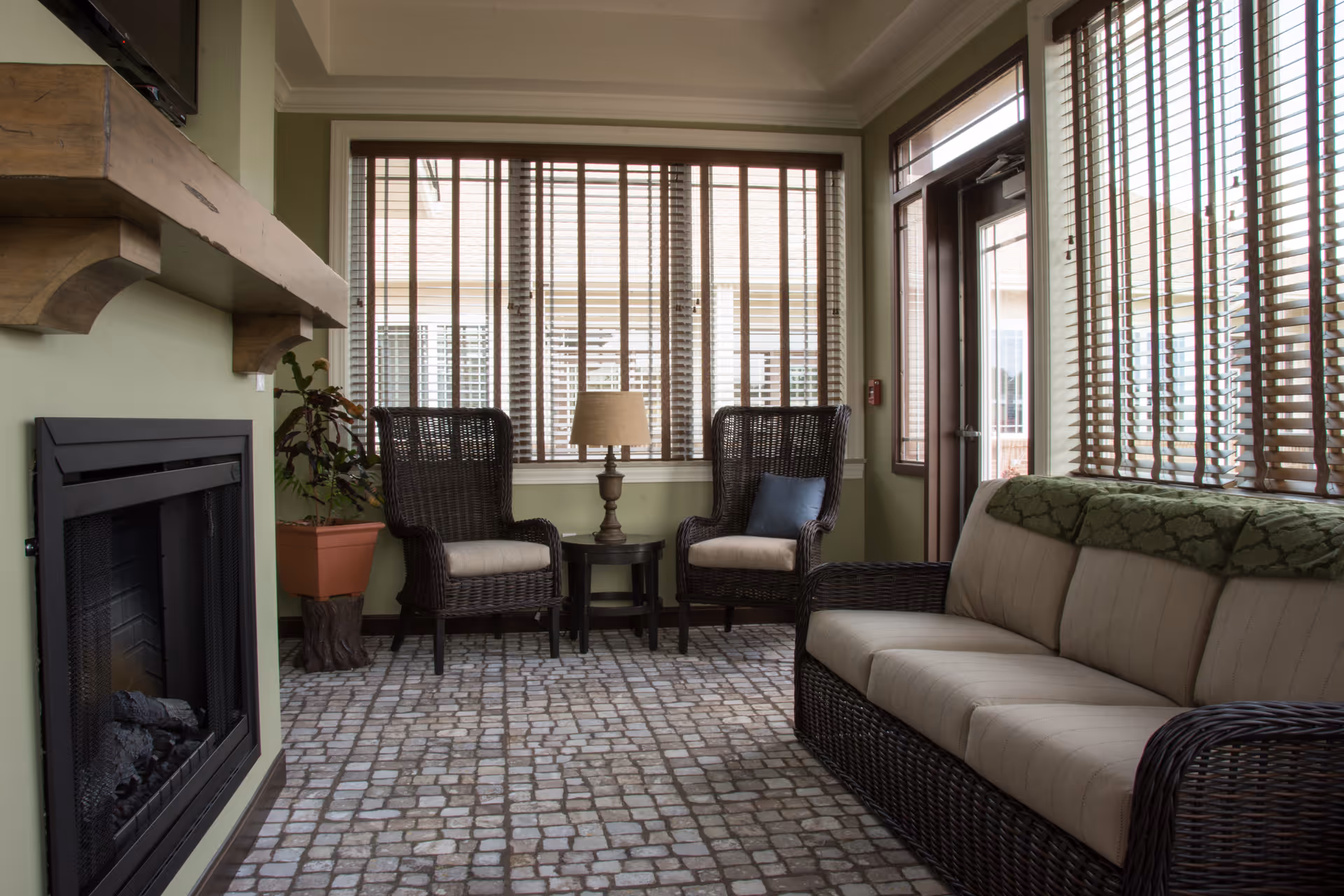 Cozy senior living lounge with wicker seating, sofa, fireplace, and large windows.