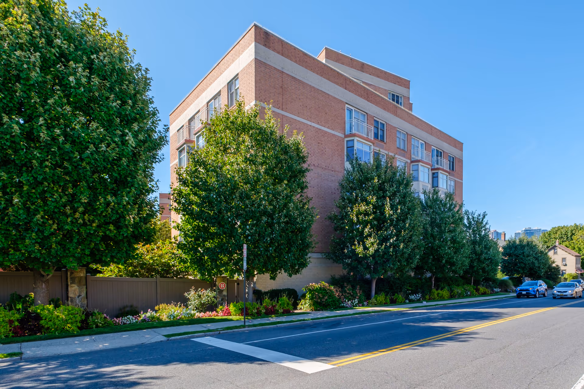 Brick multi-story residential building with trees, landscaped sidewalk and parked cars along a sunny street.