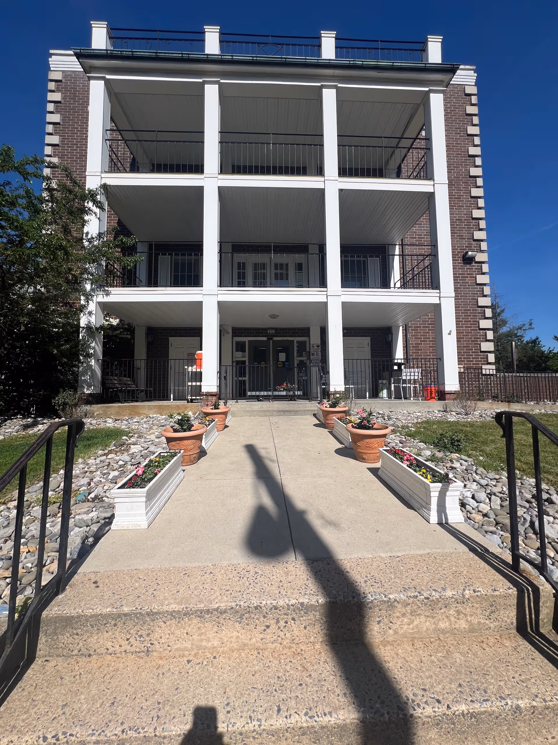 Three-story brick building with white columns, balconies, a central entrance, and planters along the walkway.