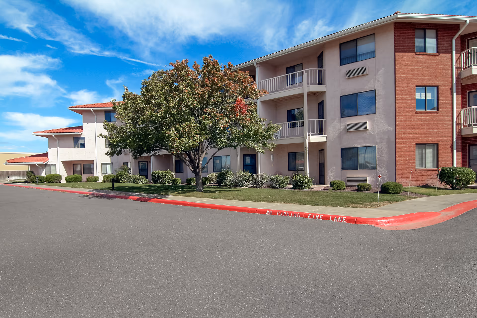 Exterior view of a multi-story residential building with balconies, surrounded by a well-maintained lawn, bushes, and a large tree. The sky is clear with some clouds, and a red-painted curb with 'NO PARKING FIRE LANE' is visible along the asphalt road in front of the building.
