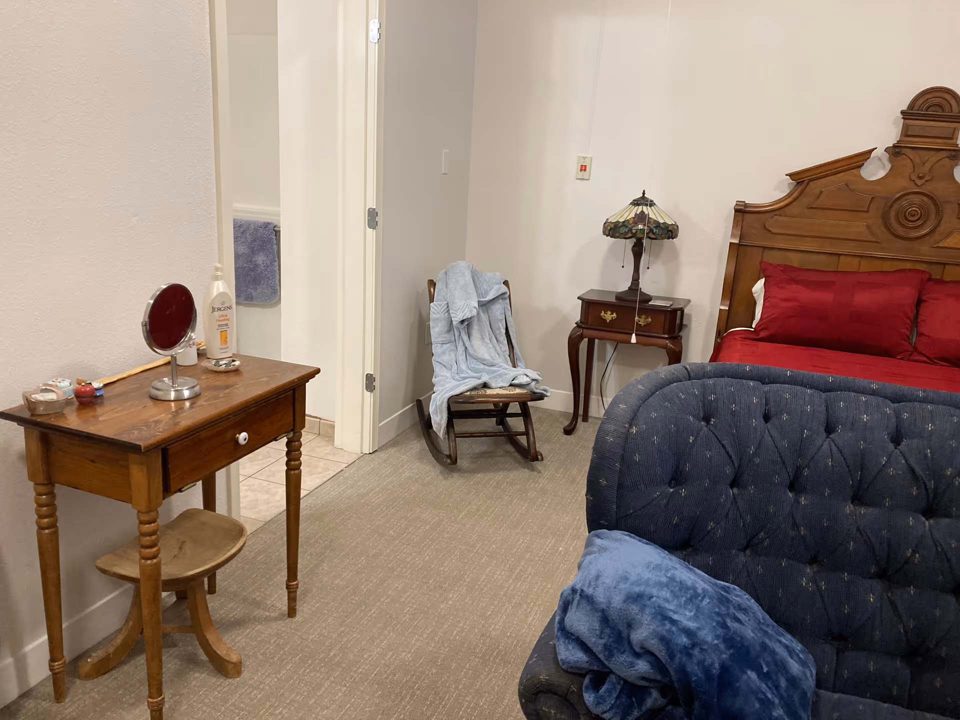 A furnished bedroom with a bed, tufted blue couch, dresser, nightstand, and doorway to a bathroom.