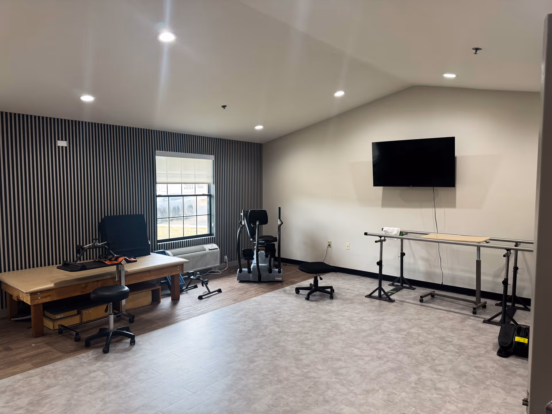 Well-lit rehabilitation therapy room with a treatment table, exercise equipment, and a wall-mounted TV.