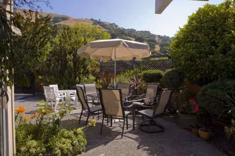 Outdoor patio with a round table, chairs, umbrella, and surrounding shrubs against hills.