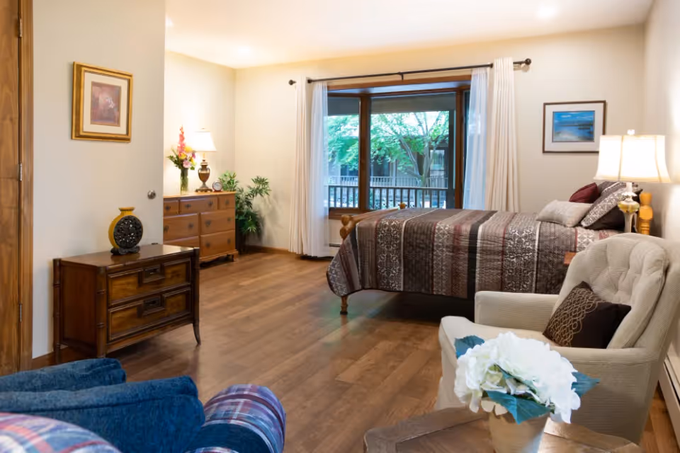 A cozy bedroom with a large window showing green trees outside. The room features a bed with a patterned brown and beige quilt, a beige armchair with a decorative pillow, a wooden dresser with a lamp and flowers, and a small wooden cabinet with a decorative vase. The floor is wooden, and the walls are light-colored with framed pictures.