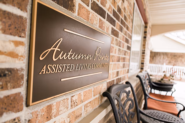 Brick exterior with a plaque reading Autumn Pointe Assisted Living Community beside a porch with chairs.