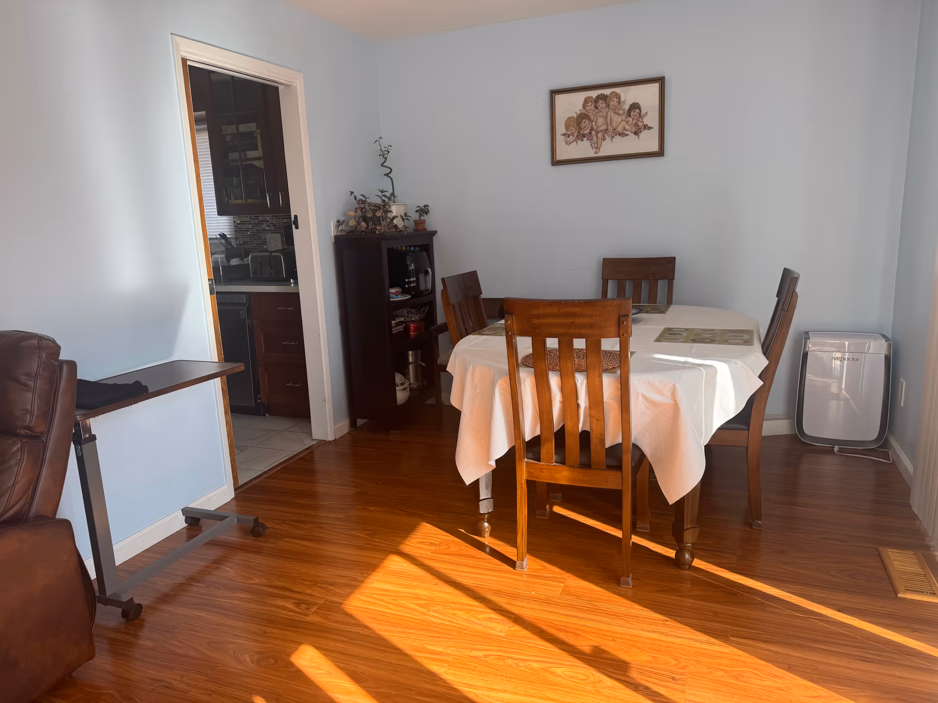 A dining area with a wooden table covered with a white tablecloth and four wooden chairs. There is a small dark wooden cabinet with plants and items on it against a light blue wall. A framed picture of cherubs hangs above the cabinet. To the left, there is an open doorway leading to a kitchen with dark cabinets. A brown leather chair and a small rolling table are partially visible on the left side. The floor is wooden, and sunlight streams in, casting shadows on the floor.