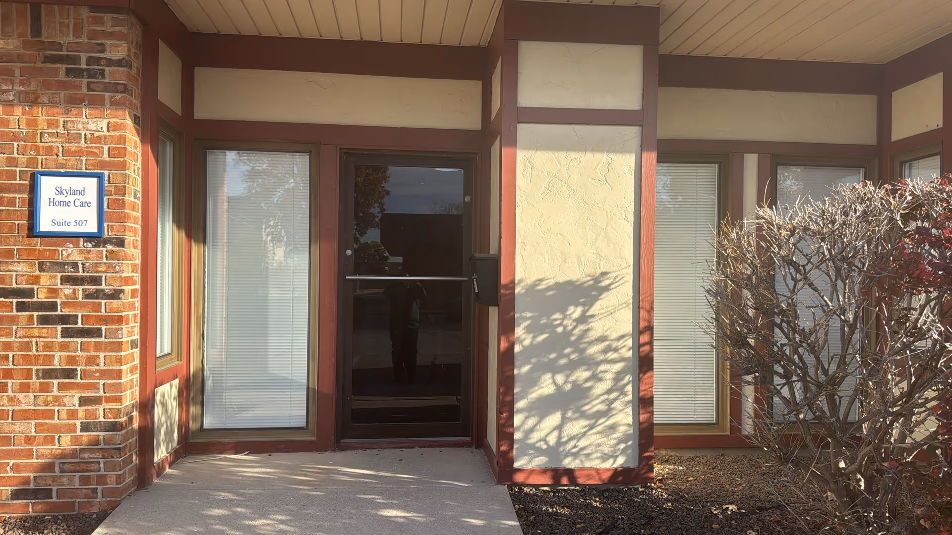 Front entrance of Skyland Home Care with glass door, brick, and shrub.