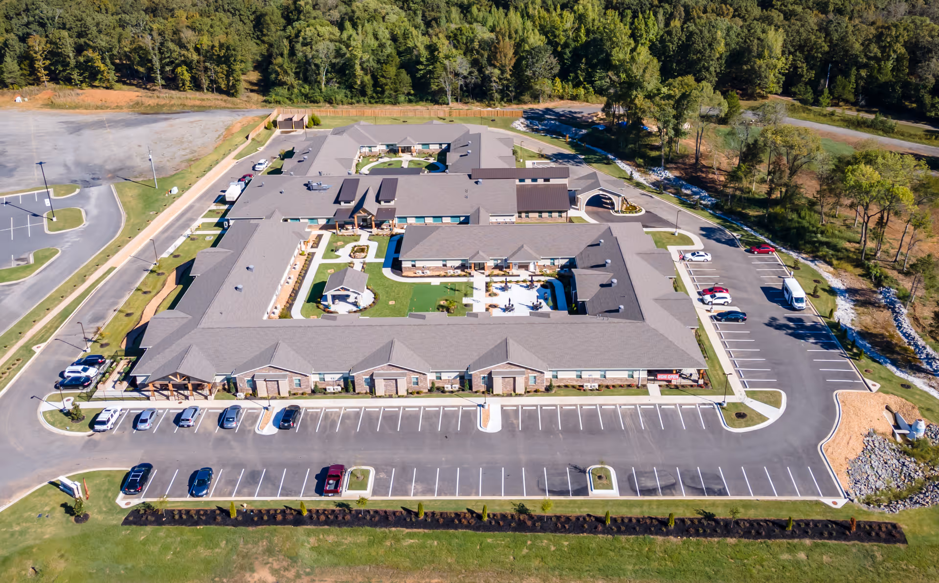 Aerial view of Crestview Senior Living facility showing a large, single-story building with a central courtyard, surrounded by parking lots and greenery. The building has a gray roof and multiple entrances, with cars parked in designated spaces around the facility.