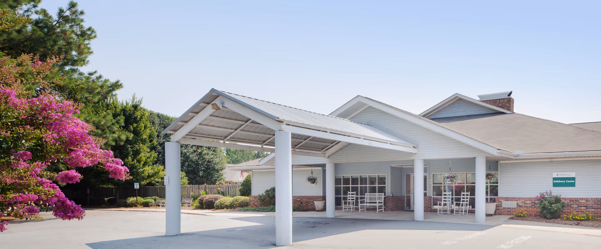 Exterior view of Salisbury Center, a single-story building with a covered entrance supported by white columns. There are rocking chairs on the porch and blooming pink flowers on a tree to the left. The sky is clear and blue.
