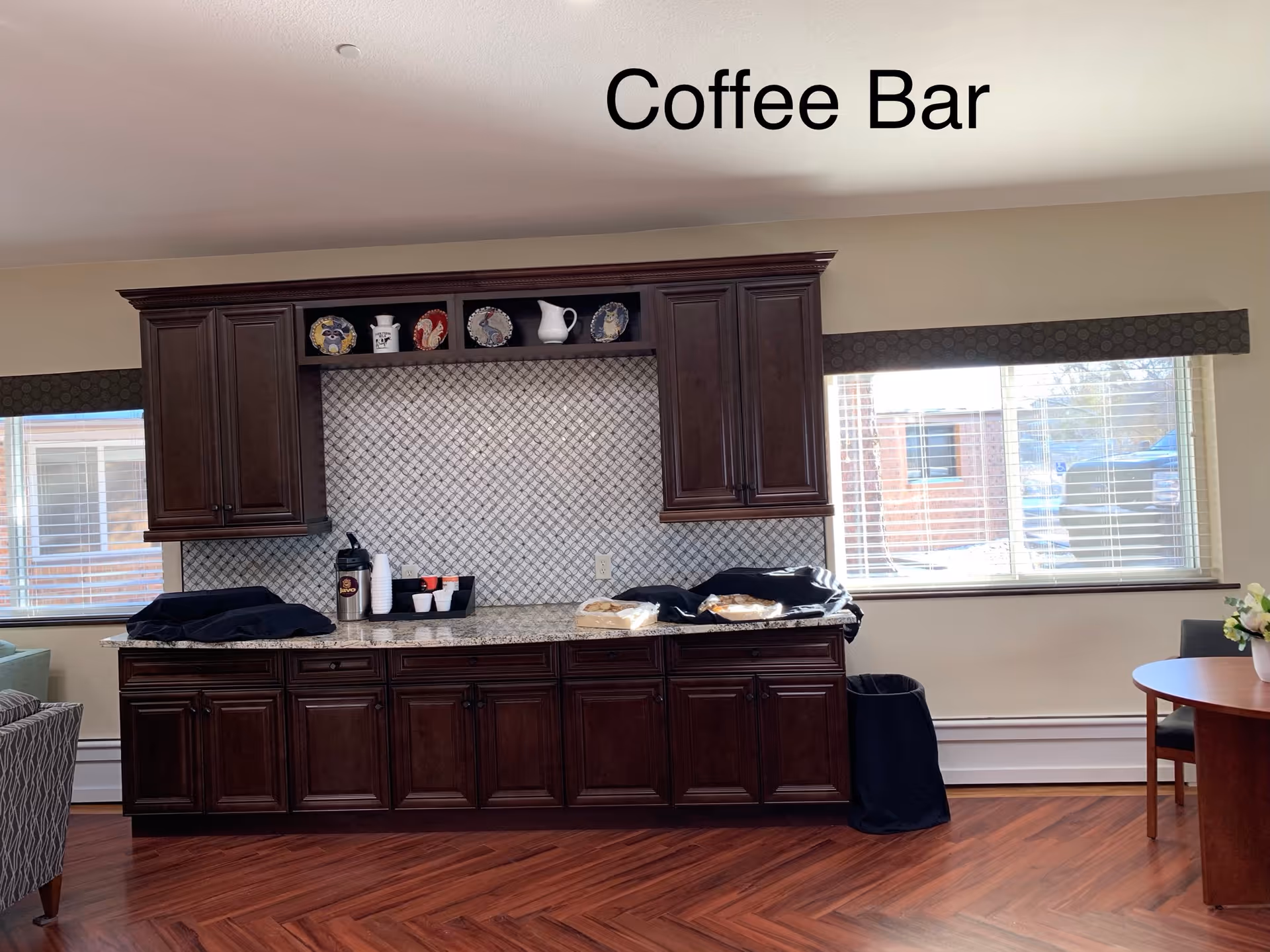 Interior view of a coffee bar area with dark wooden cabinets, a granite countertop, and a tiled backsplash. There are decorative plates and a white pitcher on the upper shelves. On the countertop, there are coffee supplies including cups and a thermos, along with some covered items. The room has large windows with blinds letting in natural light, and a round table with chairs is partially visible on the right side.