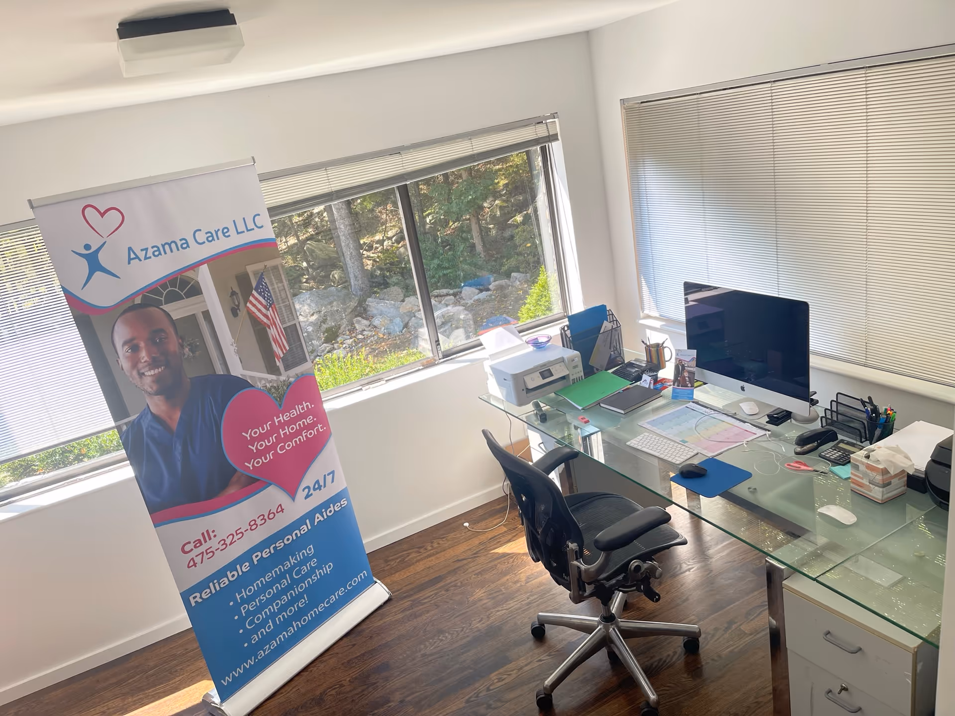 Office workspace with glass desk, computer, printer, and Azama Care LLC banner beside a sunlit window.
