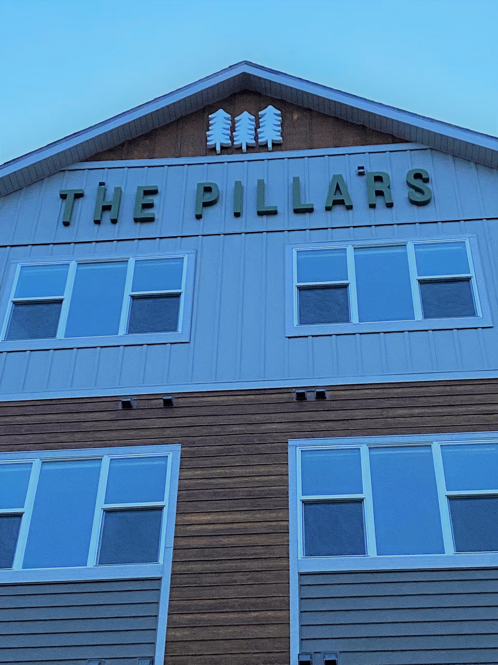 Exterior view of a multi-story building with a combination of wood and light-colored siding. The building has several windows and a sign near the roof that reads 'THE PILLARS' with three tree icons above the text.