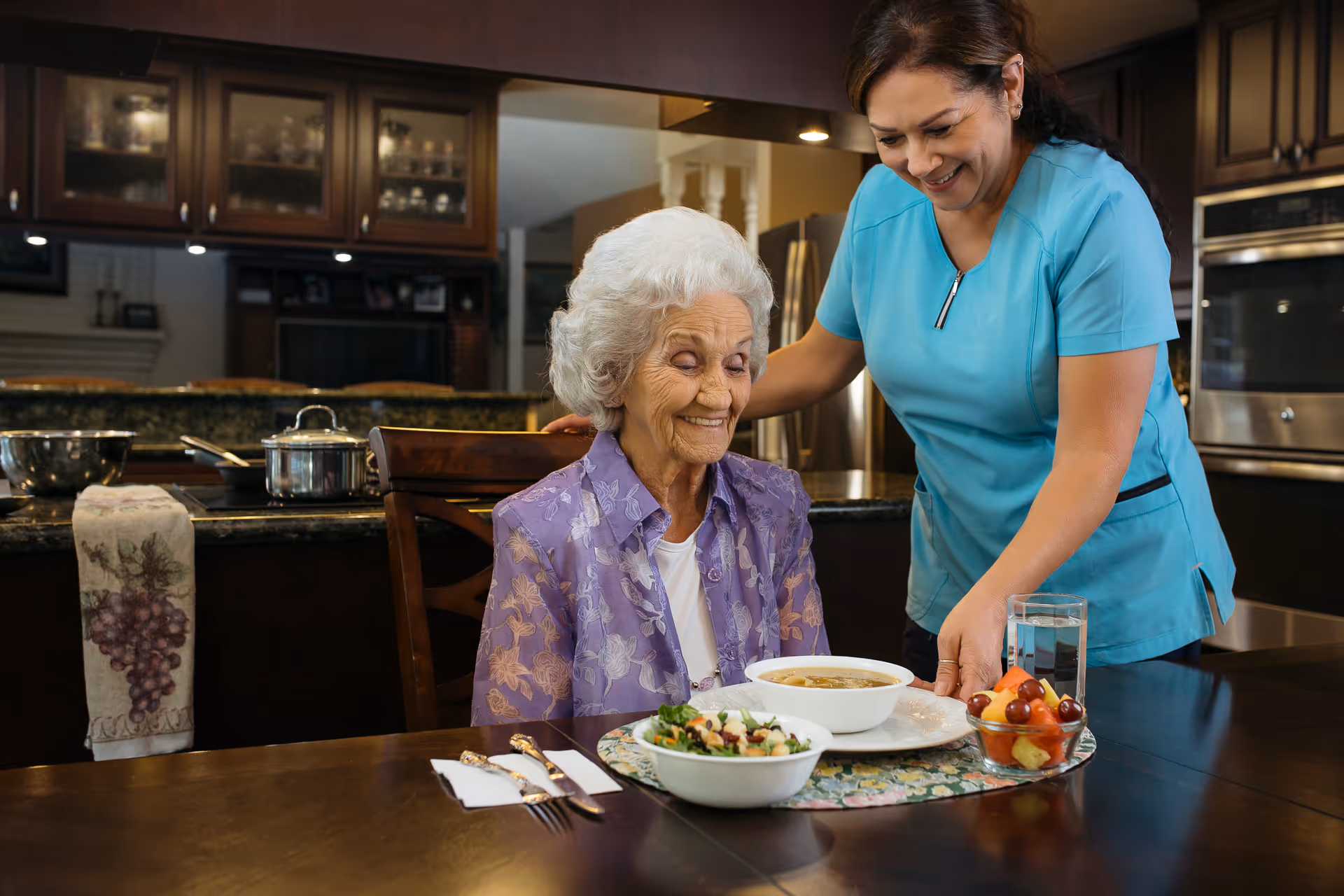 A caregiver in blue scrubs smiles while an elderly woman sits at a dining table.