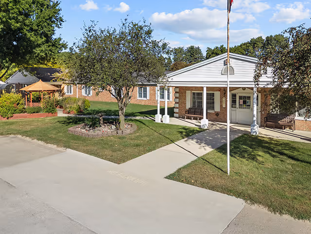 Front exterior of a single-story brick senior living facility with a covered entrance, flagpole, lawn, trees, and a gazebo.