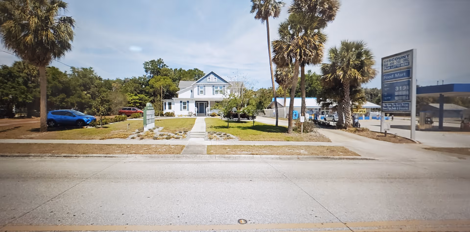 A white, house-style senior living facility with palm trees and a parking lot.