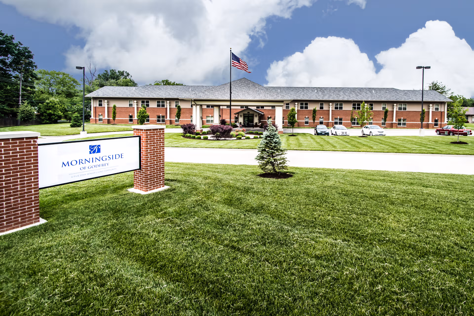 Exterior view of a brick senior living building with a manicured lawn and flagpole.