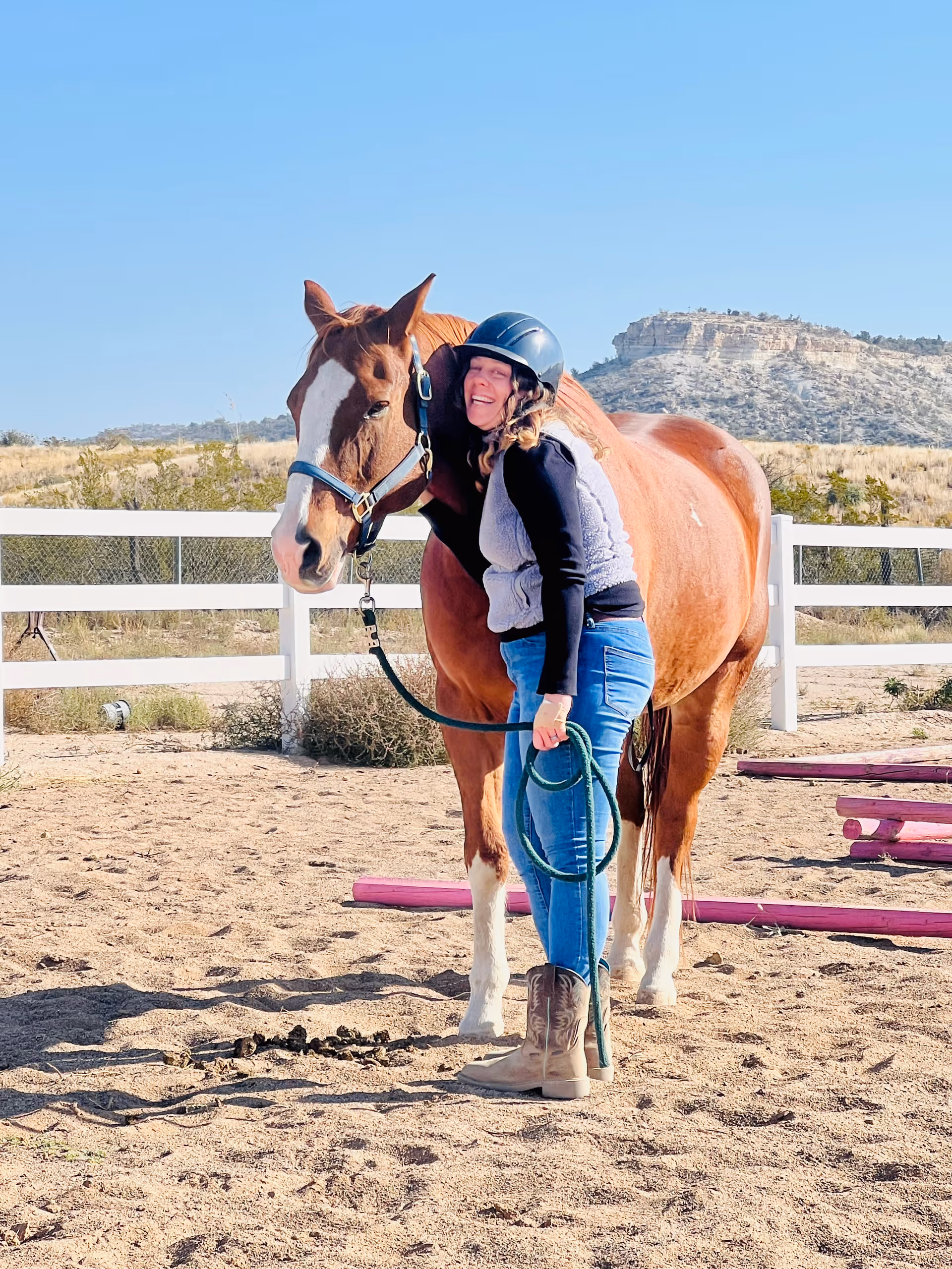 A woman wearing a helmet and casual outdoor clothing stands next to a brown horse with a white blaze on its face in a sandy fenced area. The woman is smiling and hugging the horse's neck. In the background, there are hills and clear blue sky.