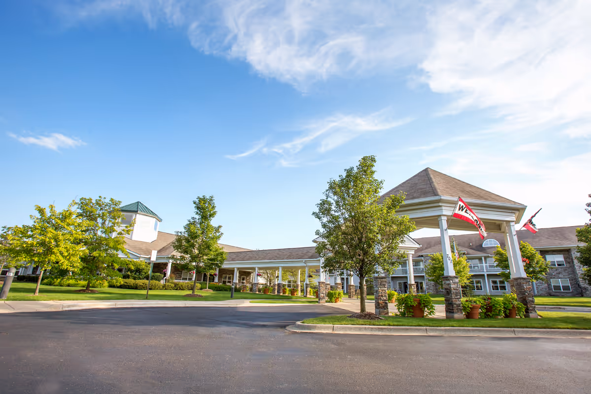 Front entrance of a senior living building with a covered porte-cochere, flags, landscaping, and a blue sky.