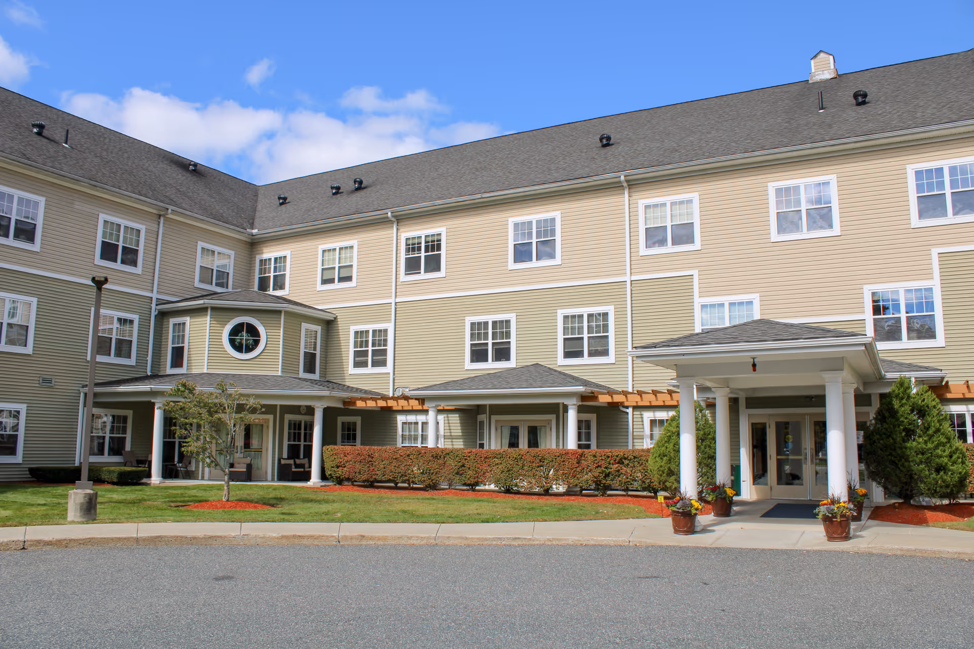 Exterior view of a multi-story assisted living facility building with beige siding, white trim, and multiple windows. The entrance features a covered porch with white columns and potted plants. There is a small landscaped area with bushes, a tree, and a lawn in front of the building under a partly cloudy sky.